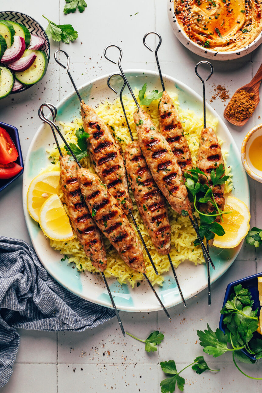 Plate of grilled chicken kefta kebabs over lemon rice with hummus and veggies in bowls around it