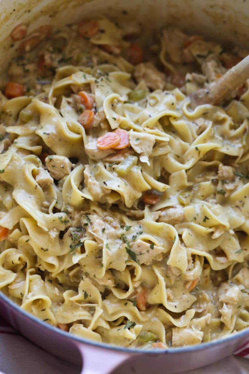 A wooden spoon stirring a large pot of chicken and noodles with carrots and seasoning