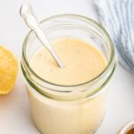 A creamy honey mustard dressing in a glass jar with a metal spoon inside the jar.