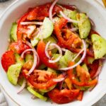 In a small white bowl, a cucumber tomato salad with sliced red onion, salt, and pepper.