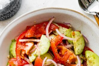 In a small white bowl, a cucumber tomato salad with sliced red onion, salt, and pepper.