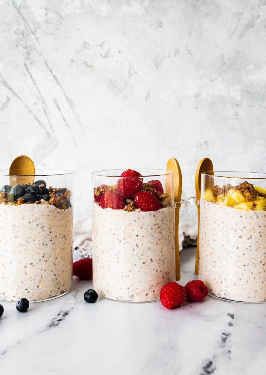 Three glass cups of kefir overnight oats. One cup is topped with fresh blueberries, another with fresh raspberries, and the last with fresh pineapple.