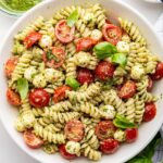 A pesto pasta salad in a large white bowl. The pasta salad has cherry tomatoes and fresh basil in it.