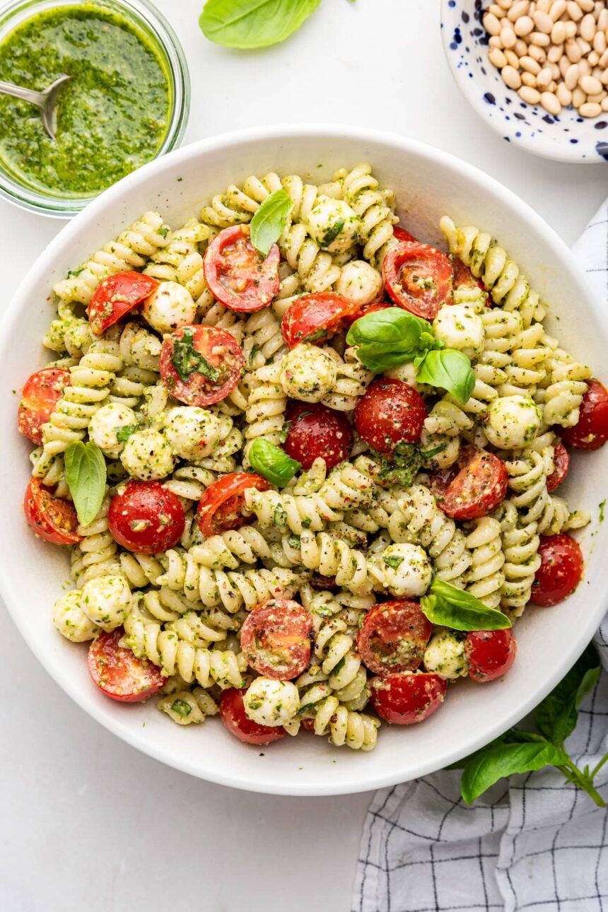 A pesto pasta salad in a large white bowl. The pasta salad has cherry tomatoes and fresh basil in it.