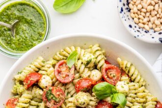 A pesto pasta salad in a large white bowl. The pasta salad has cherry tomatoes and fresh basil in it.