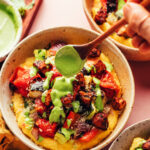 Drizzling creamy pesto sauce from a spoon onto a bowl of polenta, sheet pan ratatouille, and crispy baked tofu