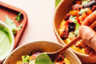 Drizzling creamy pesto sauce from a spoon onto a bowl of polenta, sheet pan ratatouille, and crispy baked tofu
