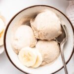 Three scoops of banana ice cream in a bowl with a metal spoon and two slices of banana.