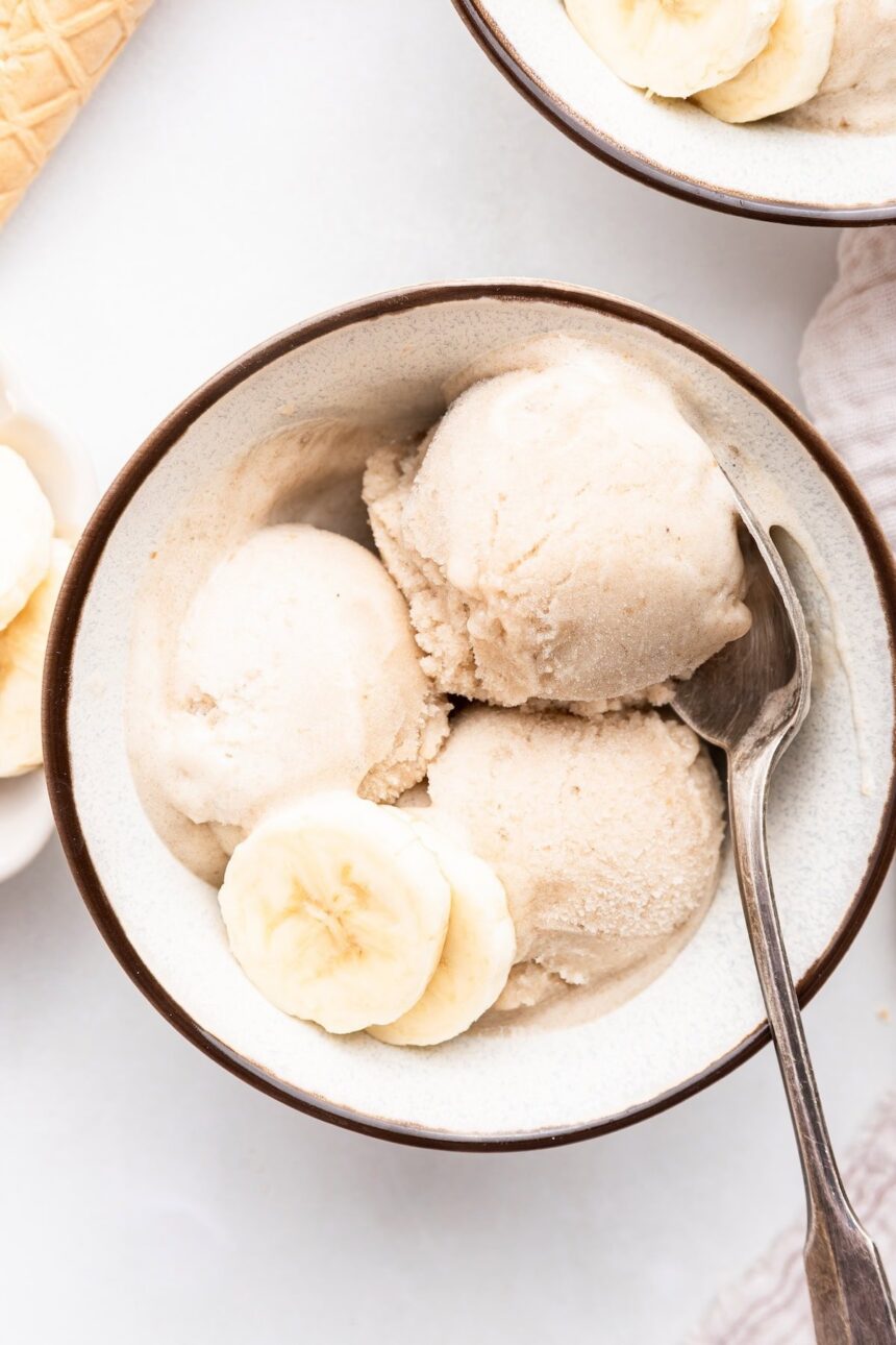 Three scoops of banana ice cream in a bowl with a metal spoon and two slices of banana.