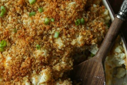 A cheesy cauliflower casserole with portions scooped out, and a wooden spoon resting in the dish.