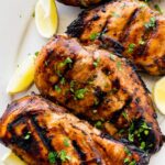 Four marinated grilled chicken breasts on a plate topped with fresh herbs and lemon wedges on the side.