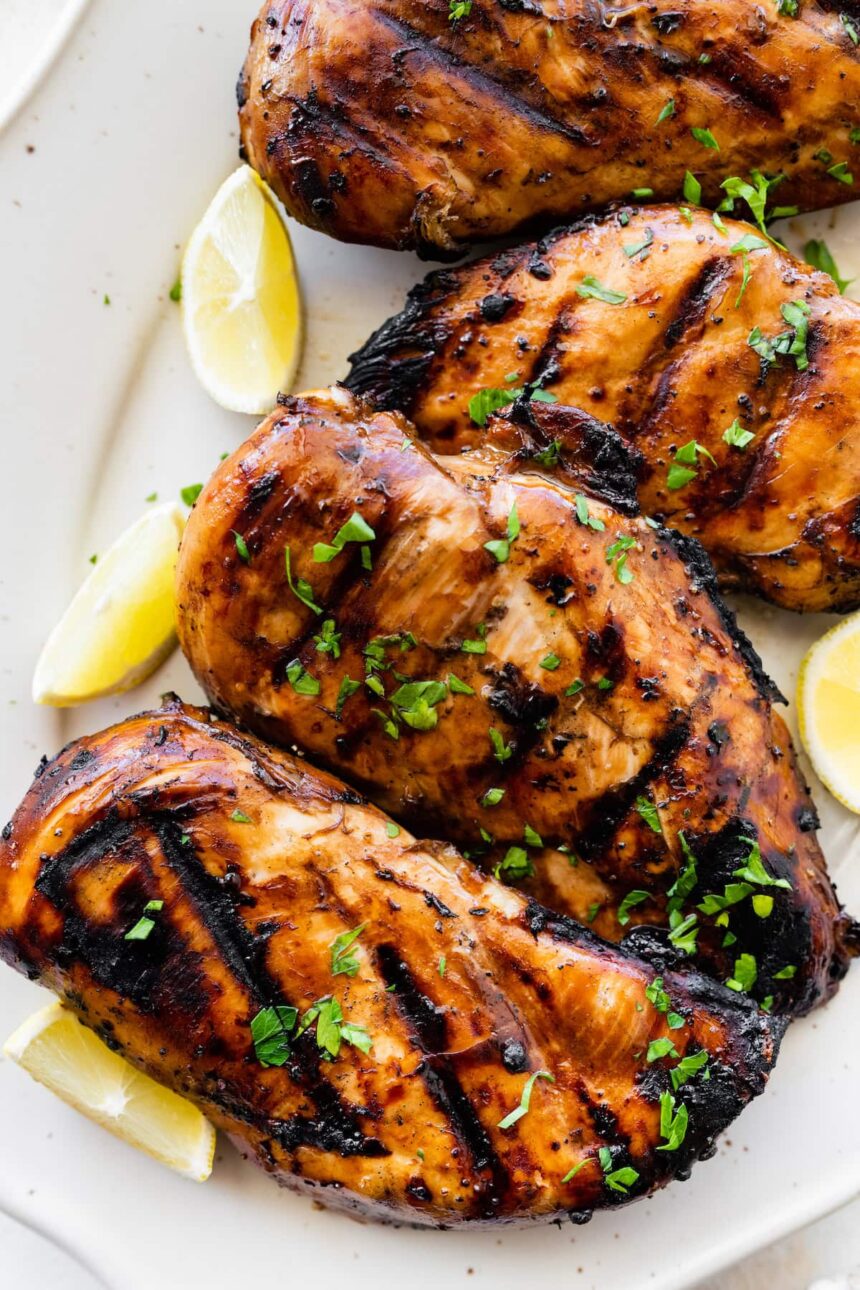 Four marinated grilled chicken breasts on a plate topped with fresh herbs and lemon wedges on the side.