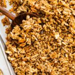 A baking sheet with a large wooden spoon taking a portion of coconut granola.