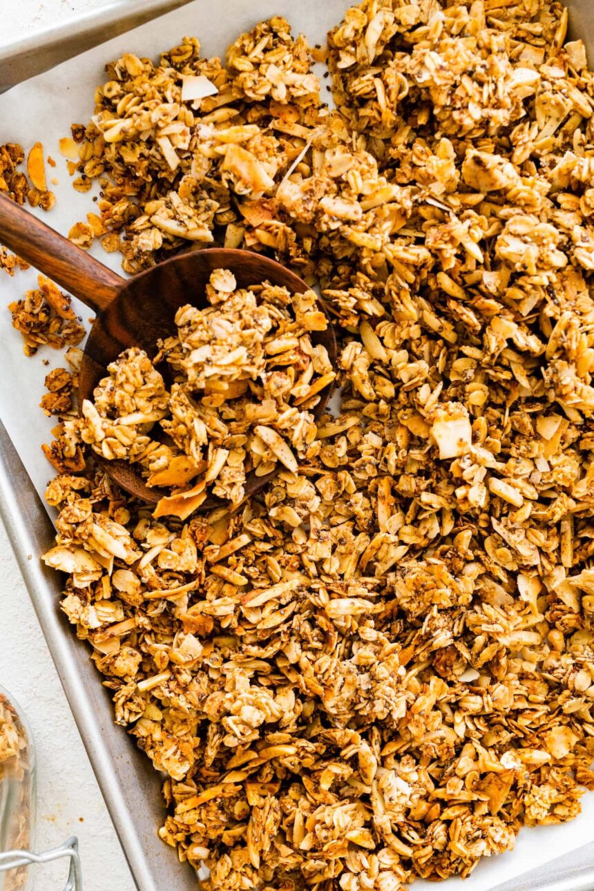 A baking sheet with a large wooden spoon taking a portion of coconut granola.