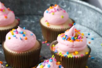 Confetti Cupcakes with pink frosting and sprinkles on top in a tray covered in sprinkles