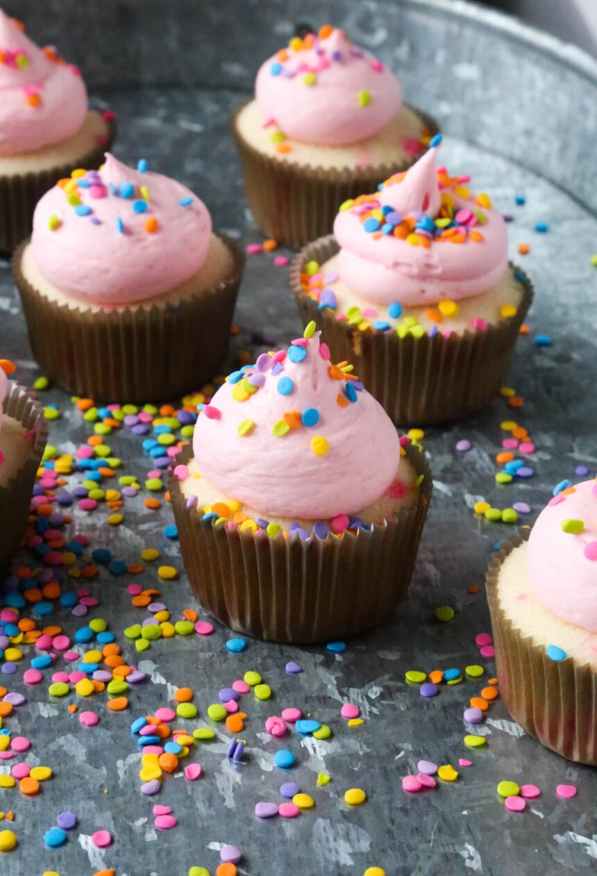 Confetti Cupcakes with pink frosting and sprinkles on top in a tray covered in sprinkles