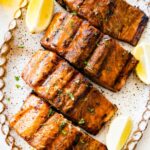 Four grilled salmon filets on a large plate with lemon wedges on the side.