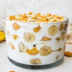 A glass trifle dish filled with healthy banana pudding. Layers of fresh banana slices and wafer cookies are visible in between the yogurt and coco whip.