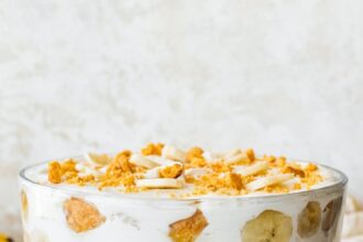 A glass trifle dish filled with healthy banana pudding. Layers of fresh banana slices and wafer cookies are visible in between the yogurt and coco whip.