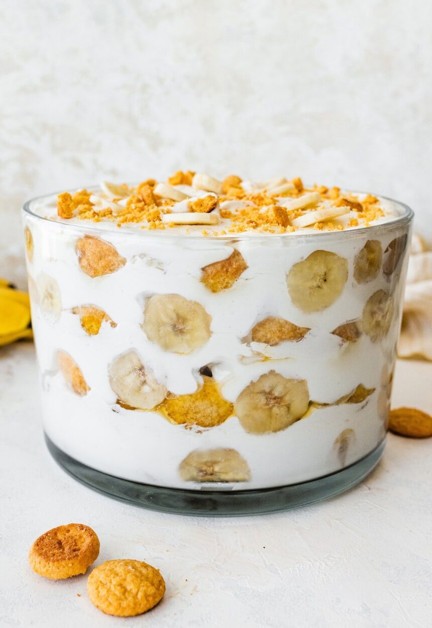 A glass trifle dish filled with healthy banana pudding. Layers of fresh banana slices and wafer cookies are visible in between the yogurt and coco whip.