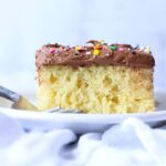 Vanilla cake with chocolate frosting and rainbow sprinkles on a white plate with a fork.