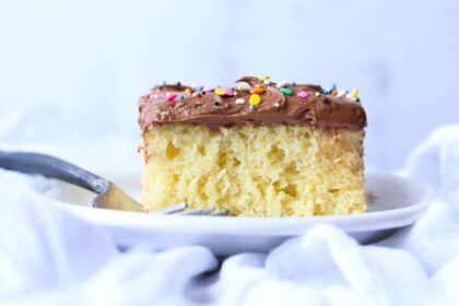 Vanilla cake with chocolate frosting and rainbow sprinkles on a white plate with a fork.