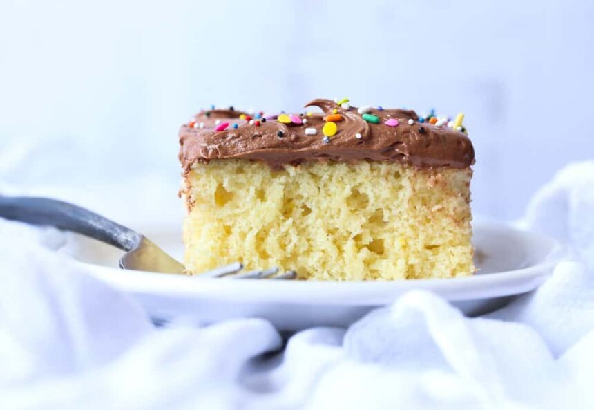 Vanilla cake with chocolate frosting and rainbow sprinkles on a white plate with a fork.