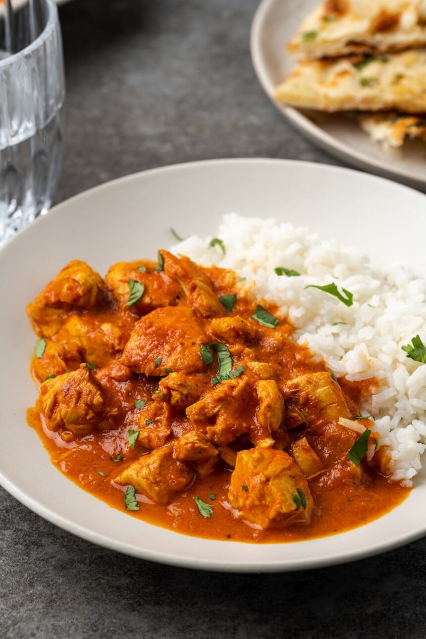 Instant pot butter chicken served on a white plate next to a side of basmati rice, garnished with fresh chopped cilantro, with a plate of naan bread in the background.