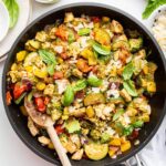 A large skillet of cooked pesto chicken with multiple vegetables and topped with fresh basil leaves.