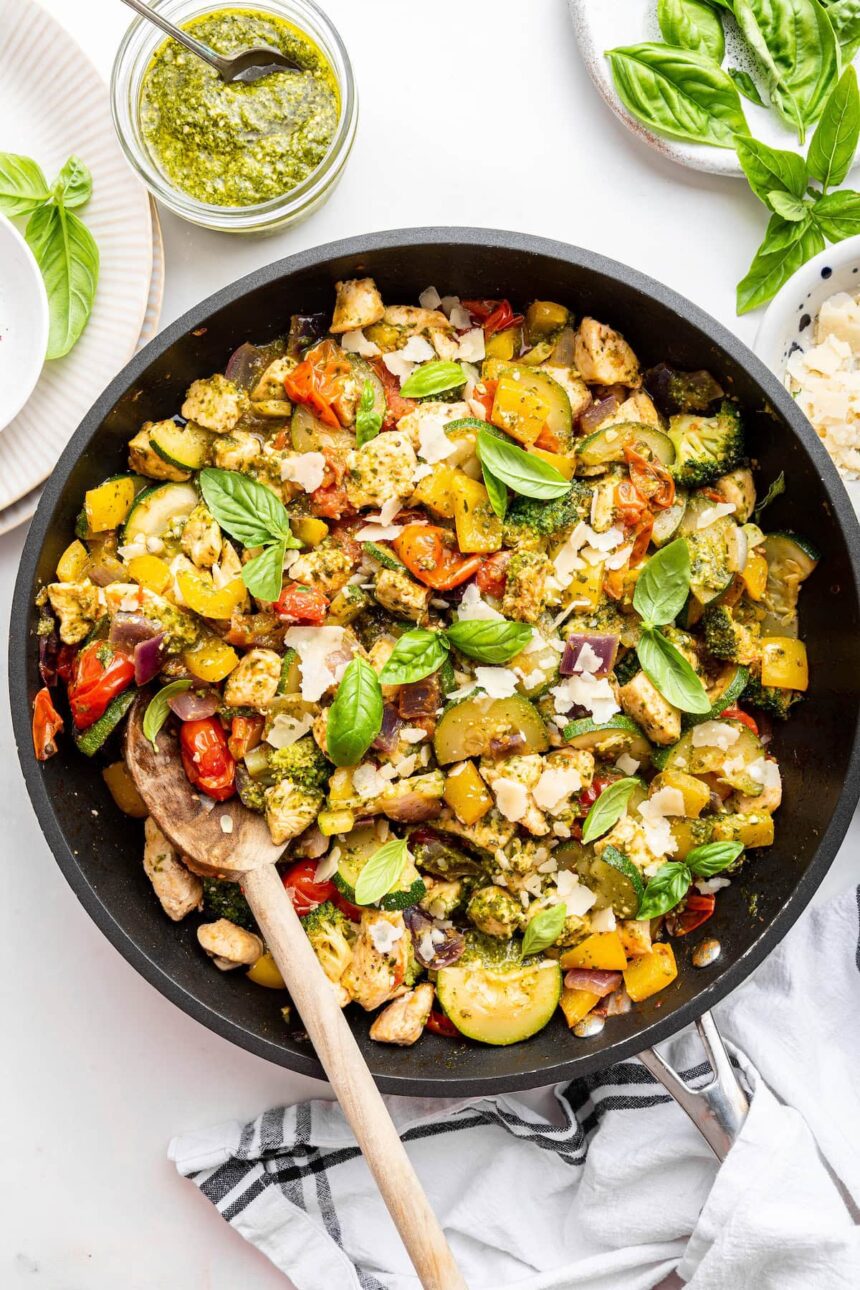 A large skillet of cooked pesto chicken with multiple vegetables and topped with fresh basil leaves.