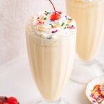 A protein milkshake in a large glass cup topped with whipped cream, sprinkles, and a cherry.