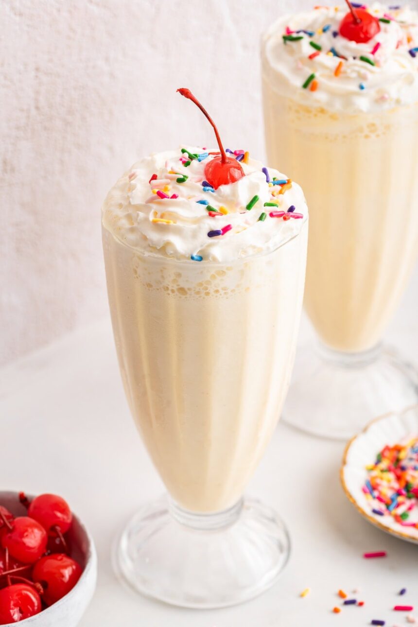 A protein milkshake in a large glass cup topped with whipped cream, sprinkles, and a cherry.