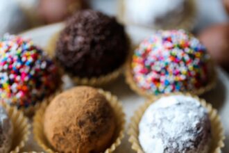 Decorated holiday rum balls in candy cups, arranged on a plate, one with a bite missing.