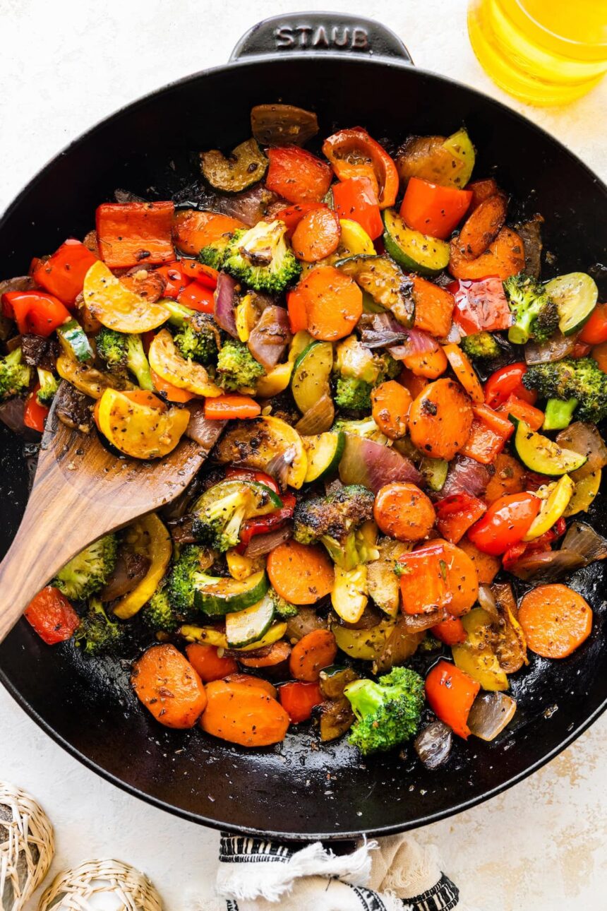 A cast iron skillet of sautéed vegetables including broccoli, carrots, onion, tomatoes, zucchini, and more.