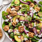 A bowl of spinach salad with ingredients like red onion, walnut, avocado, and cranberries.