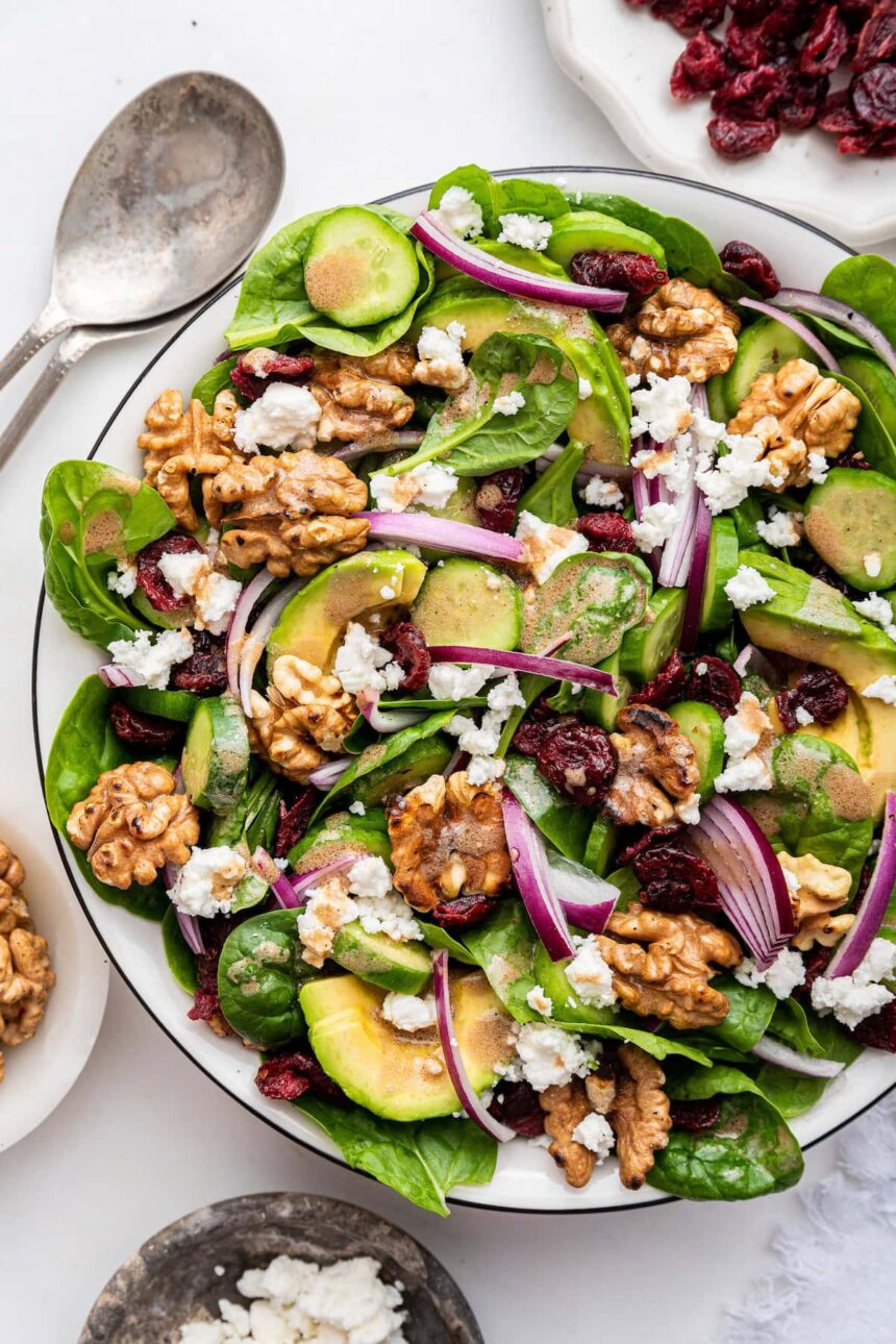 A bowl of spinach salad with ingredients like red onion, walnut, avocado, and cranberries.