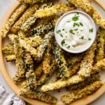 A large plate full of crispy zucchini fries covered in panko bread crumbs with a small dish of a dip.