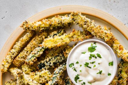 A large plate full of crispy zucchini fries covered in panko bread crumbs with a small dish of a dip.