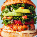 Close up photo of a salmon burger on a plate with mayonnaise, sweet chili sauce, tomato, kale apple slaw, and avocado