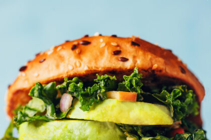 Close up photo of a salmon burger on a plate with mayonnaise, sweet chili sauce, tomato, kale apple slaw, and avocado