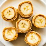 Six banana sushi roll-ups on a plate. The bananas are wrapped in a tortilla with a nut butter inside.