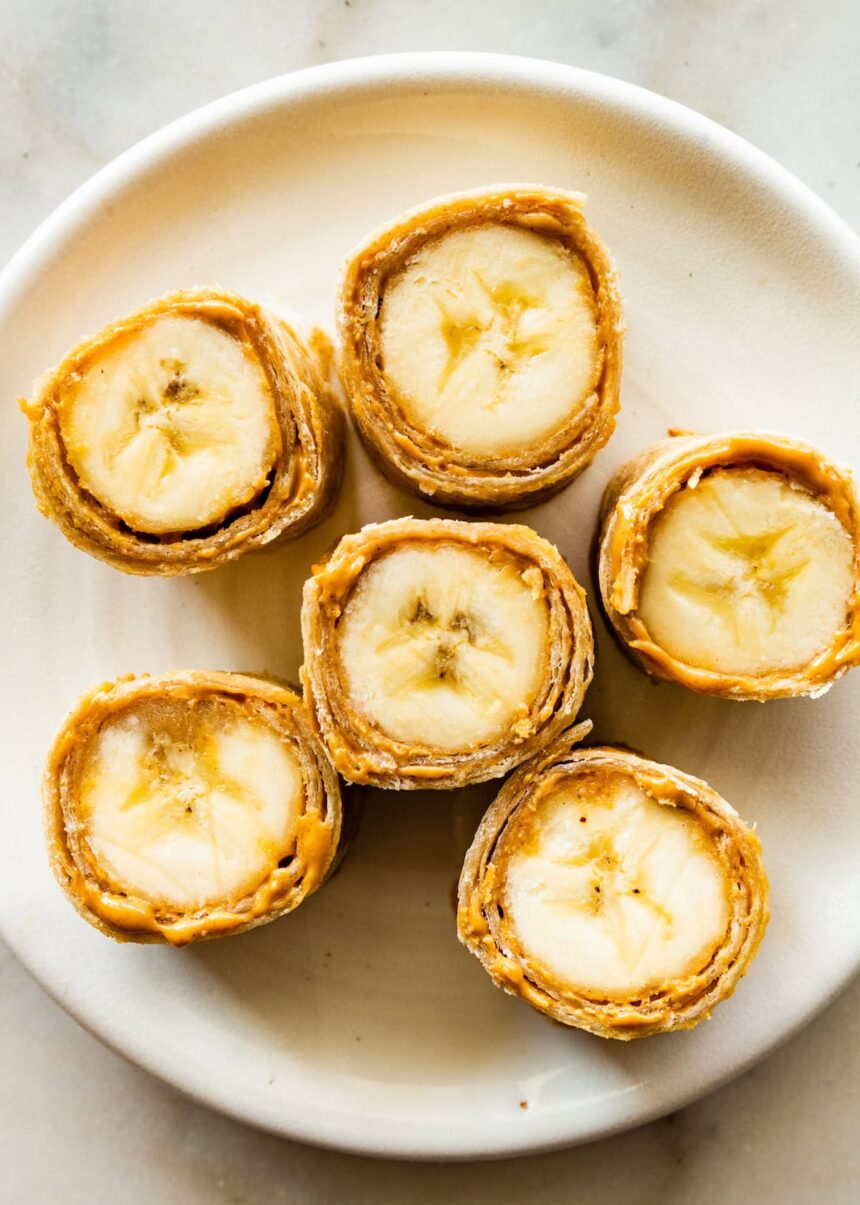 Six banana sushi roll-ups on a plate. The bananas are wrapped in a tortilla with a nut butter inside.