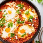 A cast iron skillet with shakshuka. It contains poached eggs in a tomato based sauce and is topped with crumbled feta cheese and fresh herbs.