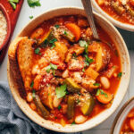 Bowls of veggie white bean minestrone topped with vegan parmesan cheese and parsley and served with toasted bread