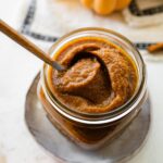 A glass jar full of pumpkin butter with a spoon inside.