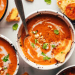 Bowls of spicy pumpkin tomato soup topped with coconut milk, cashews, cilantro, and naan