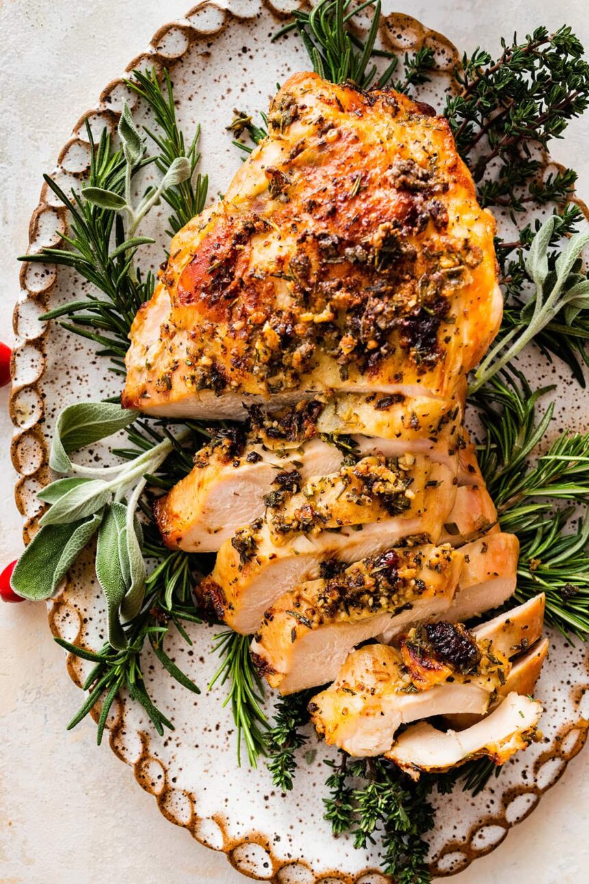 An air fryer turkey breast sliced sitting on a bed of fresh herbs and cranberries on a white platter.