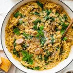 A white Dutch oven with creamy orzo, chicken sausage and spinach mixed together. A brown serving spoon is in the pot on the right hand side.