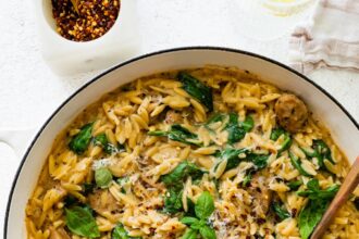 A white Dutch oven with creamy orzo, chicken sausage and spinach mixed together. A brown serving spoon is in the pot on the right hand side.