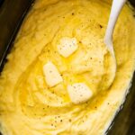 Mashed potatoes in a slow cooker with slices of butter, seasonings, and a spatula.